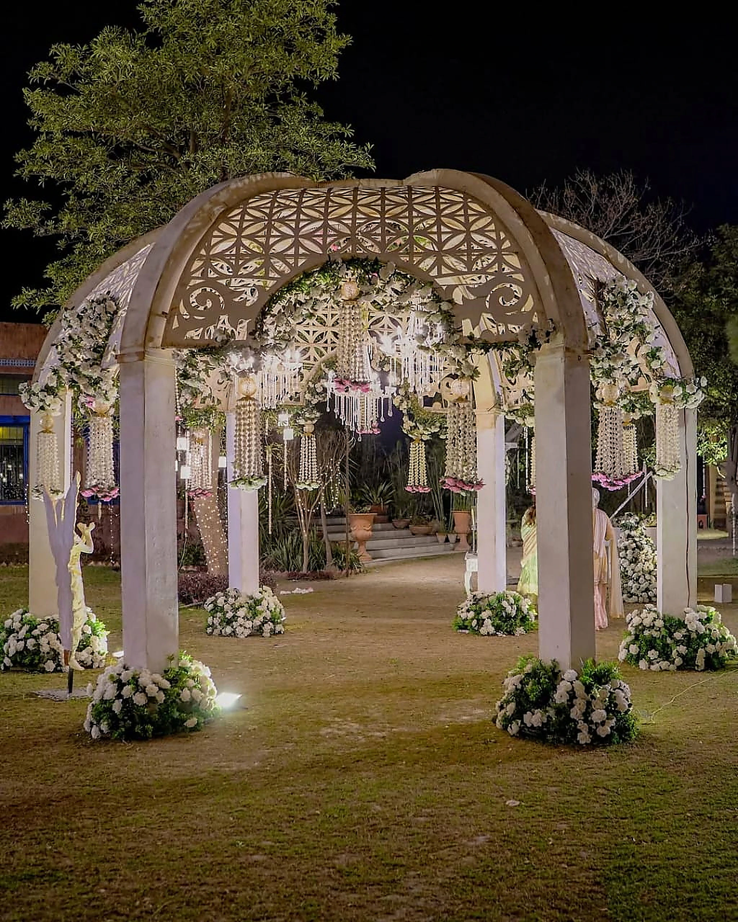 Illuminated latticed nikah pavilion framed by white florals on an evening lawn