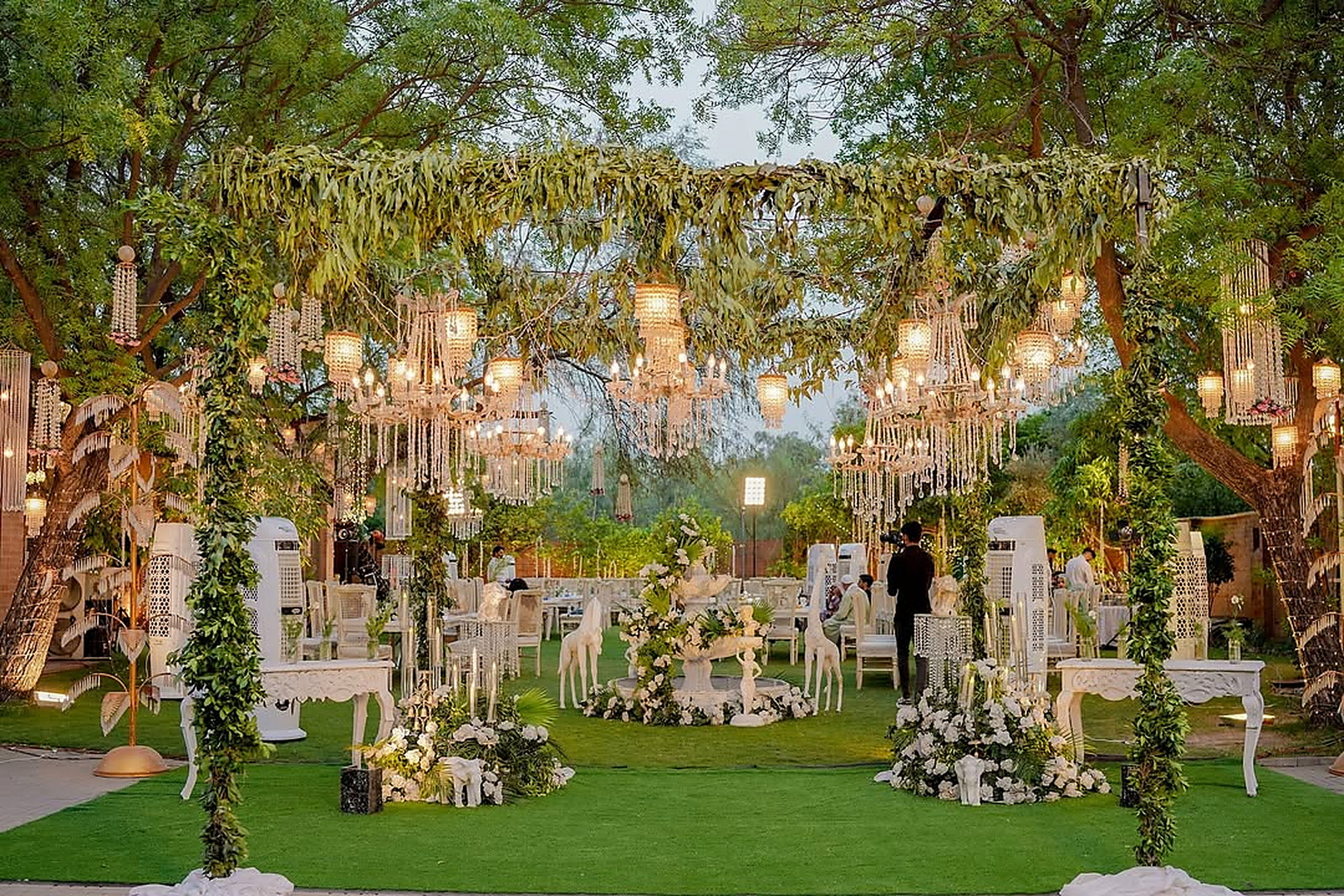 Chandeliers suspended from mature trees above a garden ceremony set